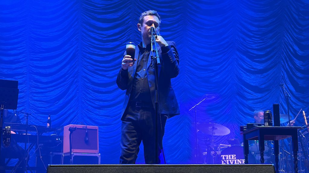 Neil Hannon in Brüssel am 17. März 2026 auf der Bühne: singend mit Glas Guinness-Bier in der Hand