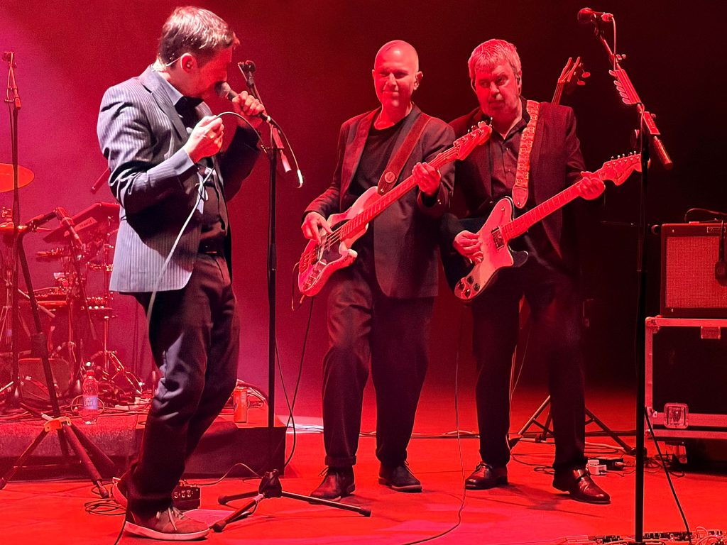 Neil Hannon auf der Bühne links von zwei Gitarristen, zunächst der Bassist, dann der Lead-Gitarrist, allesamt rot beleuchtet